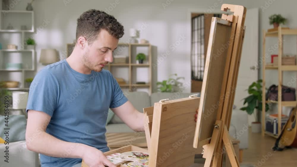 Young man is working in his modern home studio in the morning, drawing on canvas, enjoying the process of creating new artwork, inspired by natural beauty, Slow motion. Young man is working in his modern home studio in the morning, drawing on canvas, enjoying the process of creating new artwork, inspired by natural beauty, Slow motion.