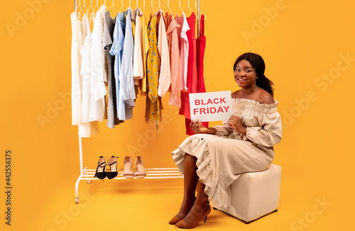 Black lady holding BLACK FRIDAY sign, offering huge seasonal sale, sitting near clothing rack on orange background