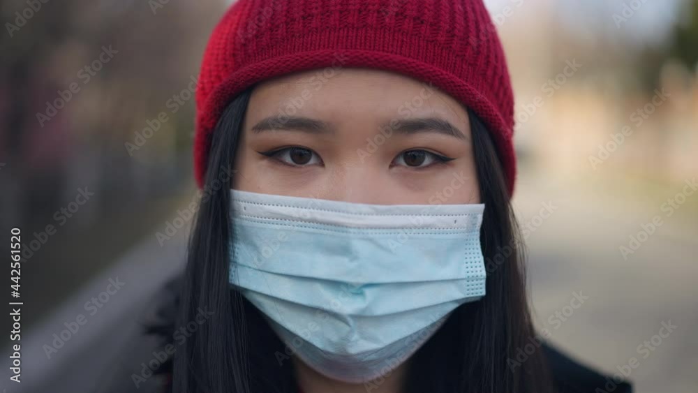 Headshot portrait of Asian young woman in Covid-19 face mask looking at camera standing outdoors. Close-up face of serious millennial posing on suburban street on coronavirus pandemic outbreak Headshot portrait of Asian young woman in Covid-19 face mask looking at camera standing outdoors. Close-up face of serious millennial posing on suburban street on coronavirus pandemic outbreak