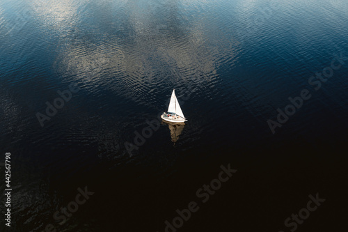 Aerial view of tiny yacht with white sails floating in calm ocean with no wind.