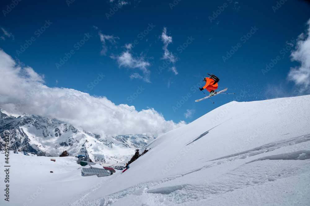 Professional athlete young male skier in an orange ski suit flies over the mountains after jumping from snow-covered ledges. Freeride sports community against the backdrop of snow-capped peaks