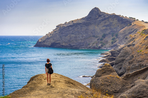 Rear view of young woman standing on mountainous road and admiring beautiful landscape of sea and mountains. Teen tourist girl walking on trail in summer day. Travel destinations, staycation concept