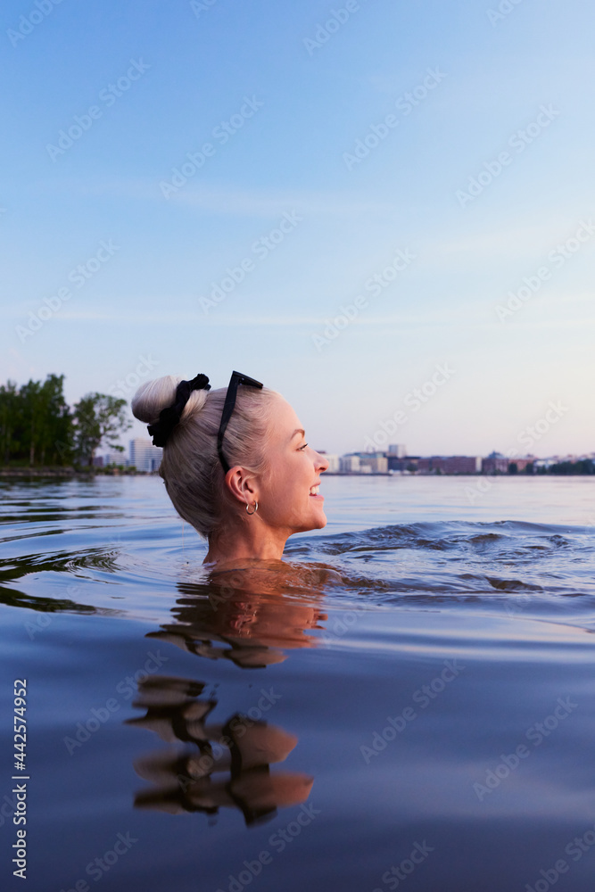 Obraz na płótnie A beautiful blond woman swimming in sea in helsinki during evening twilight sunlight sunset in nordic in finland Kalasatama in background w salonie