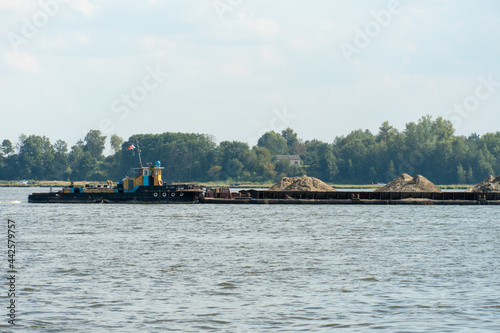 A non-self-propelled barge platform transports mineral and construction cargo, sand, crushed stone extracted from the bottom of a lake or canal along the river. A barge with sand and a tugboat