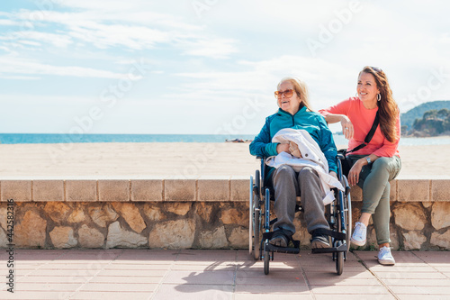Daughter sitting with aged mother in wheelchair on embankment