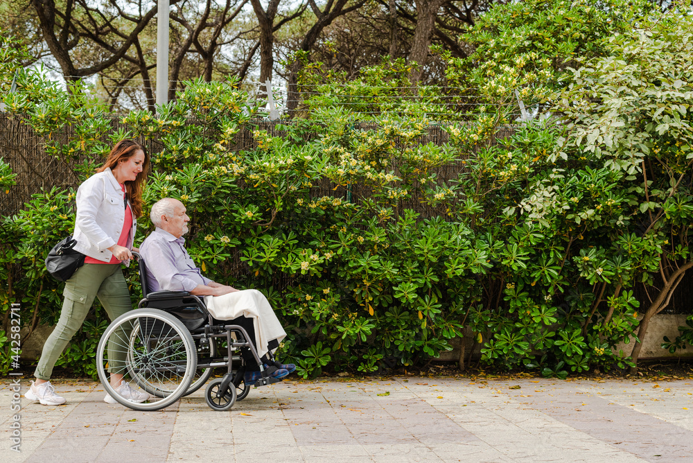 Daughter walking with elderly father in wheelchair in park Daughter walking with elderly father in wheelchair in park