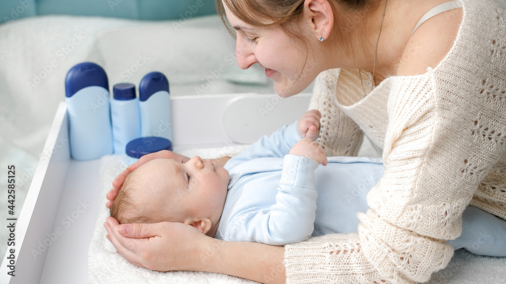 Obraz premium Smiling mother stroking and smiling at her little baby boy lying on dressing table at bedroom. Concept of hygiene, baby care and healthcare