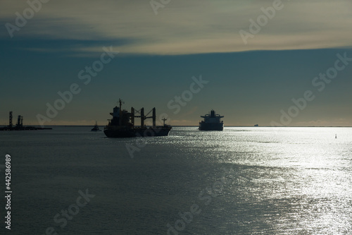 Silhouettes of cargo ships leaving the bay to the sea, accompanied by a tug, at sunset.