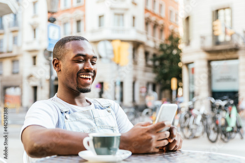 Attractive afro american man sitting at a bar terrace. He is using his phone and smiles.
