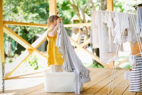 cute caucasian toddler girl in a basket with clean linen on the veranda, hangs up the washed, and helping children in the household