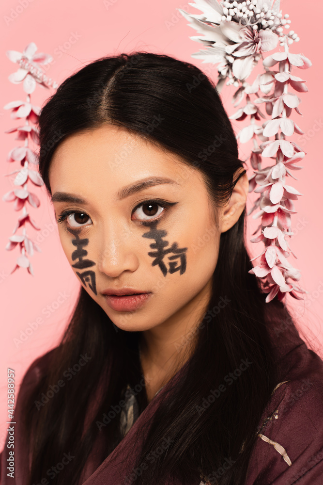 Young asian woman with hieroglyphs on face looking at camera isolated on pink Young asian woman with hieroglyphs on face looking at camera isolated on pink