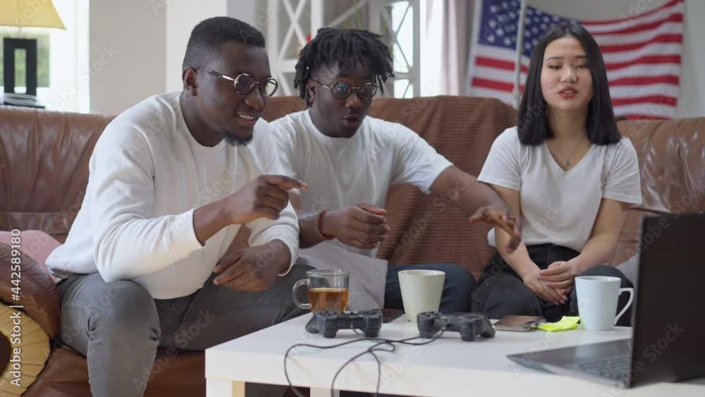 Group of young cheerful friends rejoicing win of sports team making victory gesture giving high-five laughing. Positive Asian woman and African American men watching championship on laptop