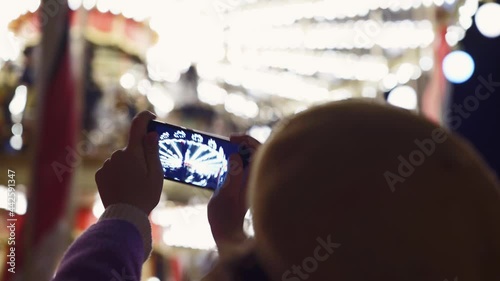 Positive young woman with curly hair in coat and beret taking photo on smartphone against glowing marry-go-round with riding people in amusement park at night photo