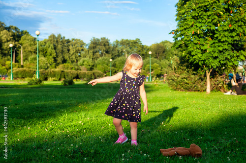 Cute little blond baby girl two year old playing with teddy bear on fresh green grass with flowers. Kid having fun making first steps on mowed natural lawn. Happy and healthy childhood concept photo