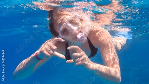Young woman in a mask with a snorkel swimming underwater, smiles and shows a heart sign with her hands photo