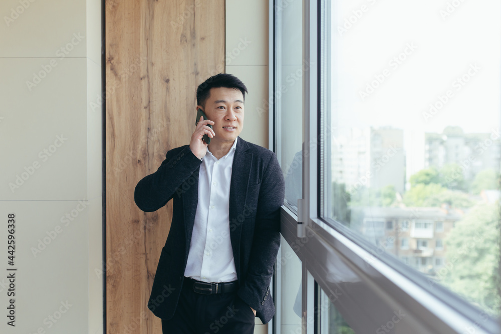 Happy and smiling Asian businessman talking on the phone in a modern office center, sharing good news with companions Happy and smiling Asian businessman talking on the phone in a modern office center, sharing good news with companions