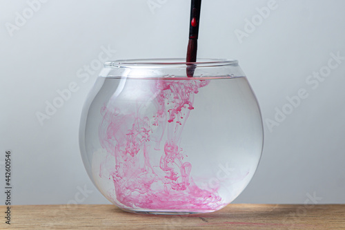 A paintbrush with red paint is dipped in a jar of water on a white background. The paint spreads in the water.