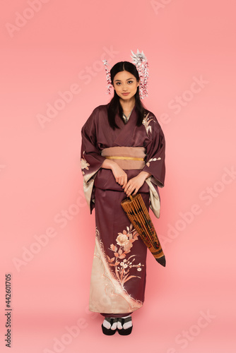 Fotografia Japanese woman with traditional hairdo and kimono holding umbrella on pink backg