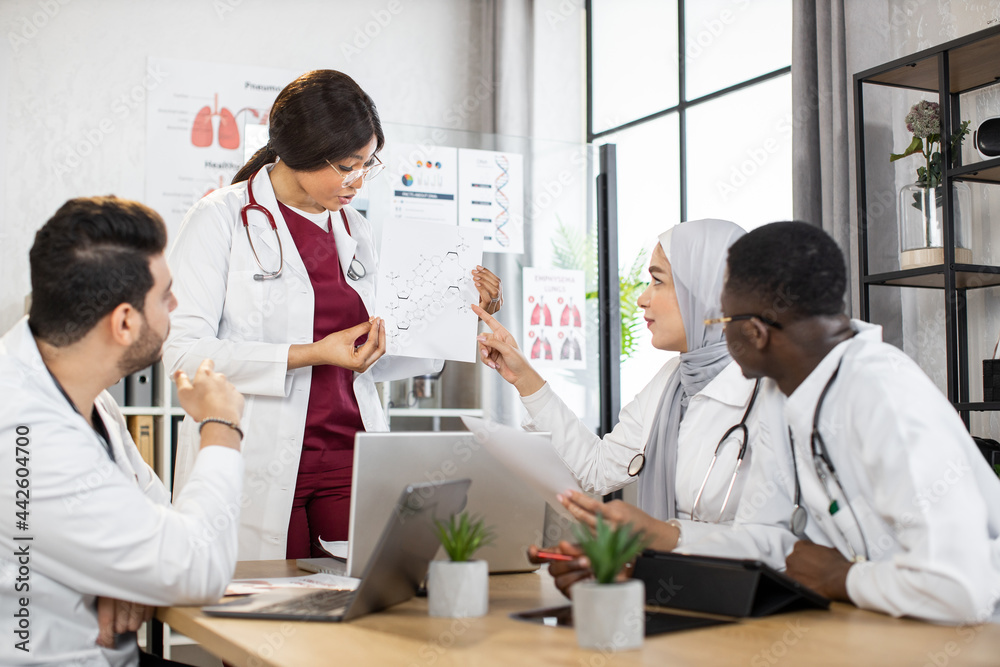 Four multiracial doctors in uniform sharing with knowledges in medicine using modern gadgets during consilium at conference room. African female doctor shows chemical structure formula for colleagues Four multiracial doctors in uniform sharing with knowledges in medicine using modern gadgets during consilium at conference room. African female doctor shows chemical structure formula for colleagues