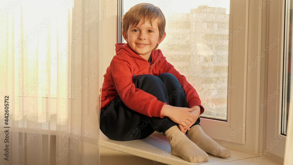 Naklejka premium Portrait of smiling little boy sitting on windowsill and looking in camera. COncept of staying at home and covid-19 pandemic