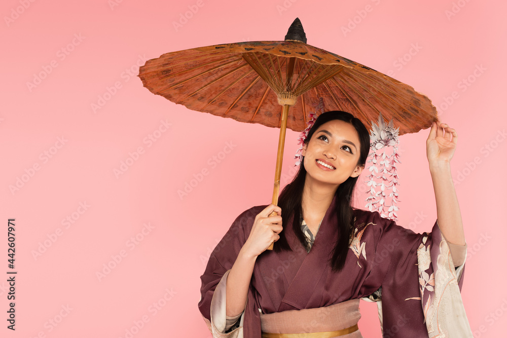 Fototapeta premium Happy japanese woman in traditional clothes standing under umbrella isolated on pink