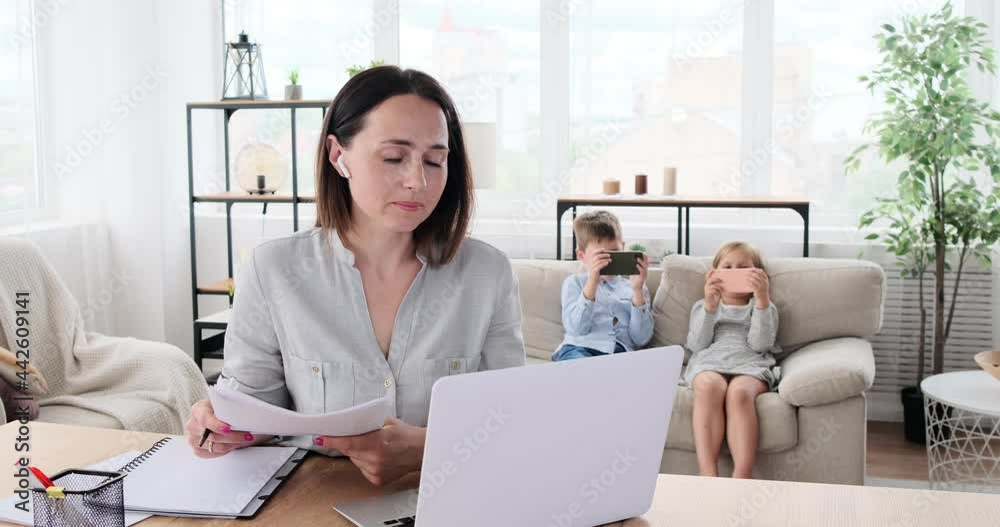 Mother doing video conference on laptop while children playing game on mobile phone in the background at home Mother doing video conference on laptop while children playing game on mobile phone in the background at home