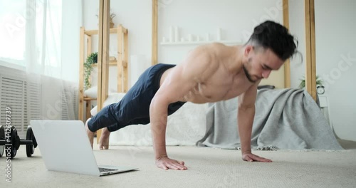 Shirtless muscular man doing push ups exercise while watching online tutorial using laptop at home photo