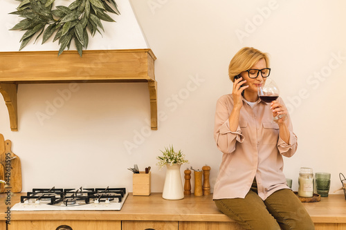 Happy mature senior elderly blonde woman in kitchen drinking red wine. Holding a glass with wine. Using mobile phone. © denis_vermenko