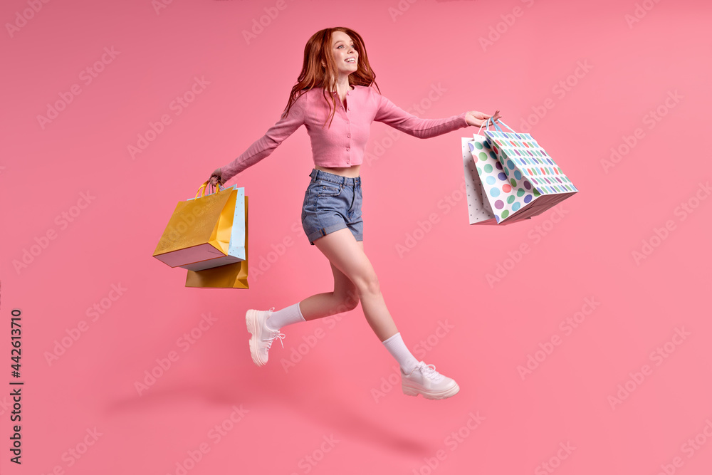 Carefree woman rejoicing with sales, shopping time, isolated on pink studio background, wearing casual outfit, denim jeans and pink shirt. portrait of jumping lady with red hair
