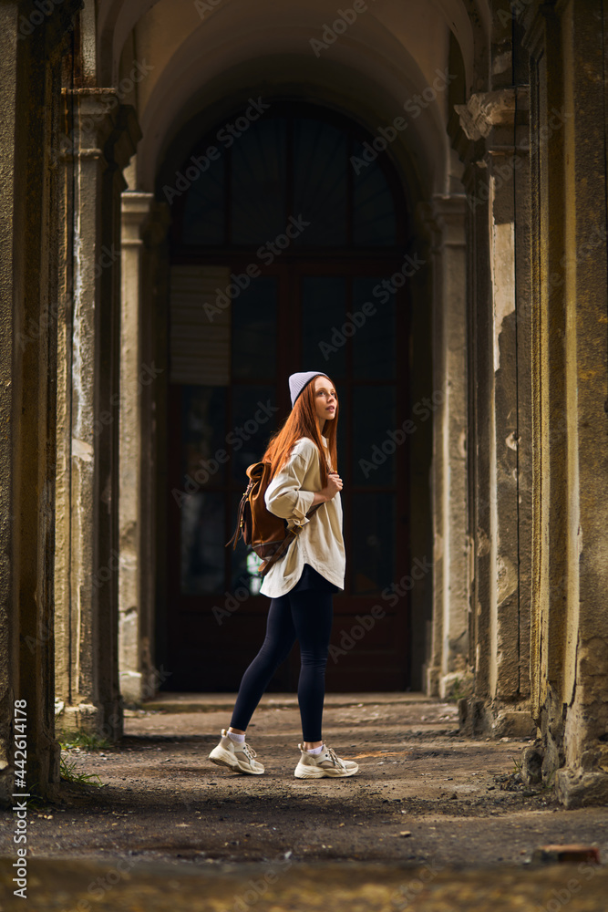 Fototapeta premium Redhead Tourist female travel destination visiting abandoned old place on vacation. Beautiful woman walking on summer holidays, cold weather outdoors. Woman is dressed in coat and hat