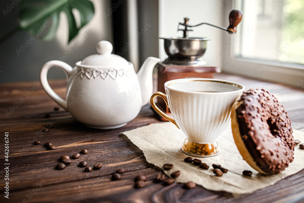 white kettle and cup on table. Breakfast by window. Chocolate donat next to scattered coffee beans. white kettle and cup on table. Breakfast by window. Chocolate donat next to scattered coffee beans.