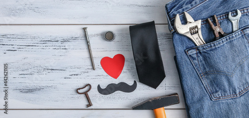 Construction tools and jeans on the table. Construction tools, jeans, a tie and a paper heart with a mustache lie on the right on a white wooden table with space for text on the left, close-up top vie © Nataliya