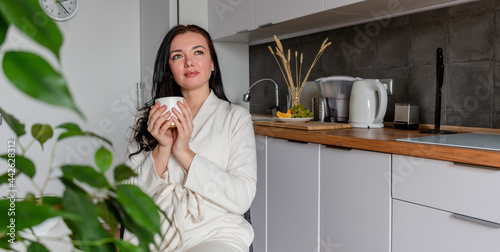Young woman with black hair in white robe sits on chair the kitchen.