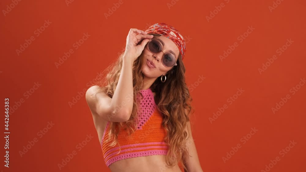 The happy curly-haired woman with bandana on her head taking off glasses and winking at the camera in the orange studio The happy curly-haired woman with bandana on her head taking off glasses and winking at the camera in the orange studio