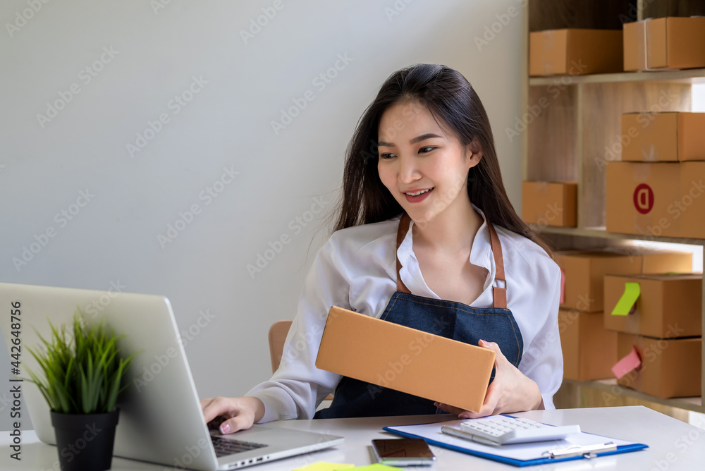 Beautiful business owner Asian wearing an apron is preparing a parcel box for delivery to a customer using a laptop at home. Beautiful business owner Asian wearing an apron is preparing a parcel box for delivery to a customer using a laptop at home.