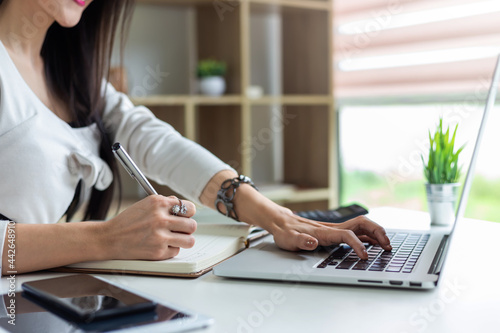 Image of a businesswoman holding a pen to taking notes while using a laptop keyboard at the office.