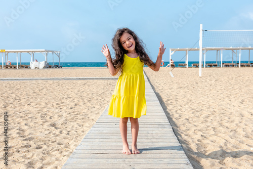 Adorable curly toddler girl in a yellow dress plays on the white sand beach 