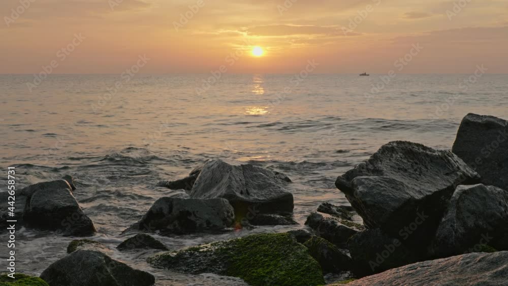 Sun rising above the sea. Waves splashing on rocks at the beach during sunrise with a boat at the background Sun rising above the sea. Waves splashing on rocks at the beach during sunrise with a boat at the background