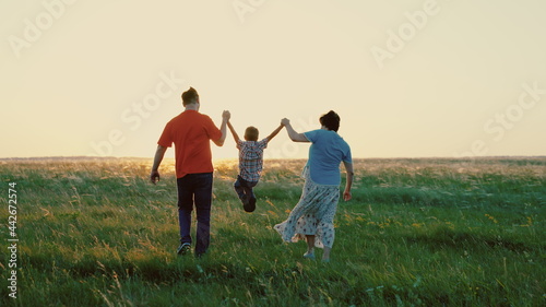 Dad  son  mom  play together run  happy child holds hands of parents  child is jumping on green grass. Family walk in park in spring at sunset  healthy childhood. Family weekend in summer in nature