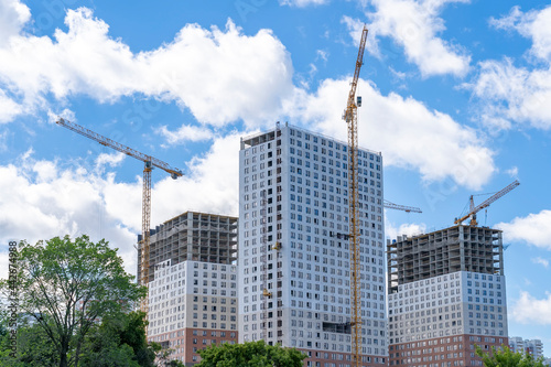 View of construction site, industrial cranes and apartment buildings at green area. Construction in city in green zone against clear blue sky. Housing construction, apartment block with scaffolding