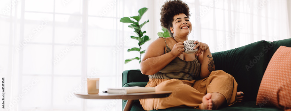Smiling woman sitting on sofa having coffee - obrazy, fototapety, plakaty 