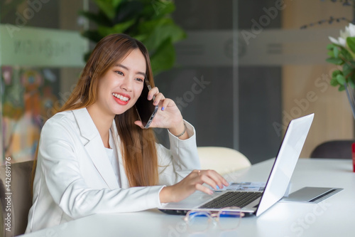 Asian beautiful working woman is calling with someone, she is talking and smiling happily while she works with her laptop in working space or office.