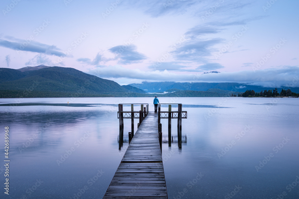Lonely woman standing at the end of Lake Te Anau jetty at sunrise, looking at the Kepler mountains. Landscape format.