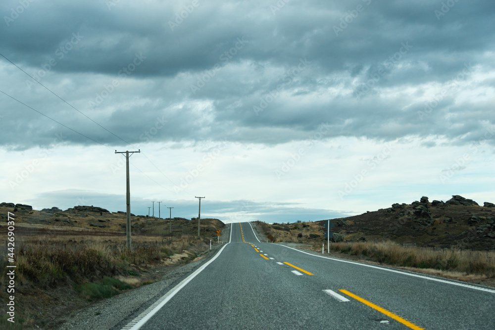 An empty country road with power lines along the road and dark clouds overhead, Otago region, South Island, New Zealand
