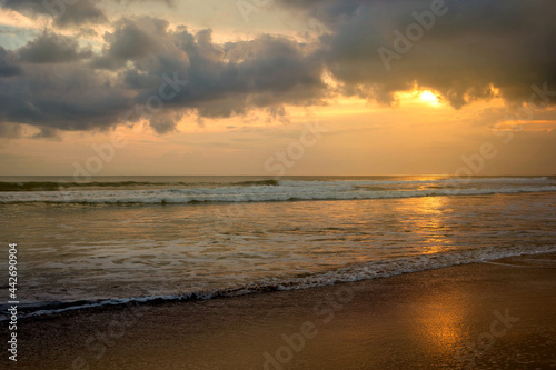 A dramatic sunset view at seaside. Orange sun reflecting in water of the ocean as tidal waves wash the sand on a shore. A beautiful natural marine landscape for wallpaper.