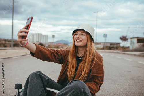 Content young lady sitting on skateboard and taking selfie in countryside photo