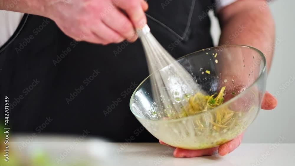 The male chef mixes the sauce with a whisk to dressing the salad in a glass bowl. Cooking in the restaurant kitchen. Close-up. The male chef mixes the sauce with a whisk to dressing the salad in a glass bowl. Cooking in the restaurant kitchen. Close-up.