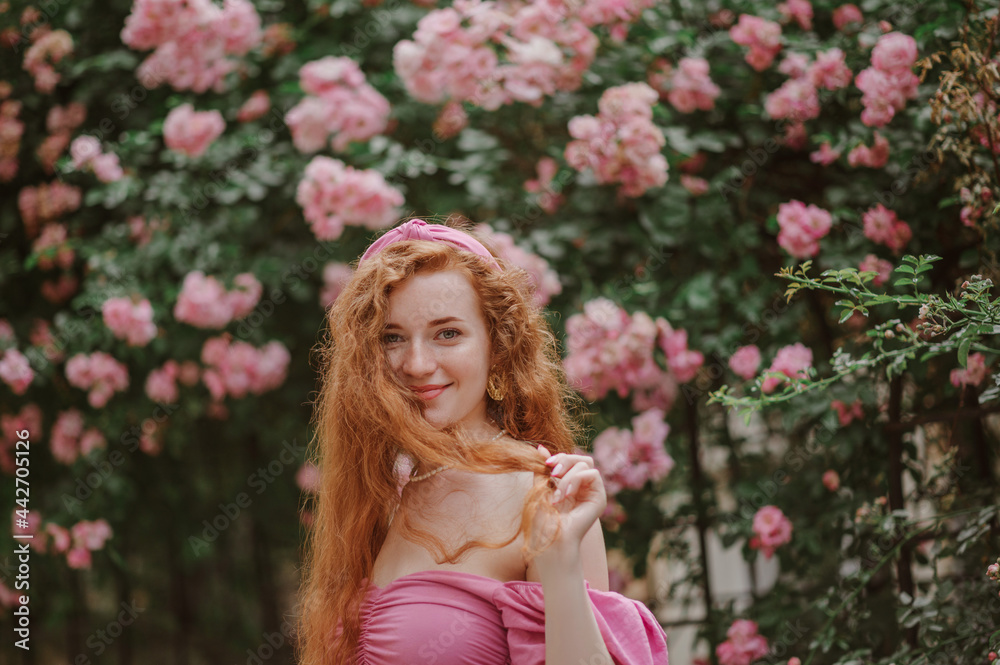 Happy smiling redhead freckled woman plays with her long natural curly hair. Model posing in blooming roses garden, wearing pink headband, blouse. Summer lifestyle, fashion, beauty concept. Copy space Happy smiling redhead freckled woman plays with her long natural curly hair. Model posing in blooming roses garden, wearing pink headband, blouse. Summer lifestyle, fashion, beauty concept. Copy space