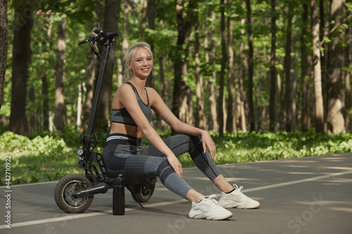A young woman is sitting on her electric scooter in the park. An electric scooter is the most environmentally friendly means of transportation. It is gaining popularity among young people