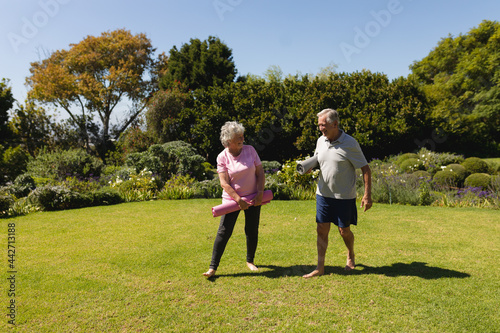Senior caucasian couple holding yoga mats and smiling in sunny garden